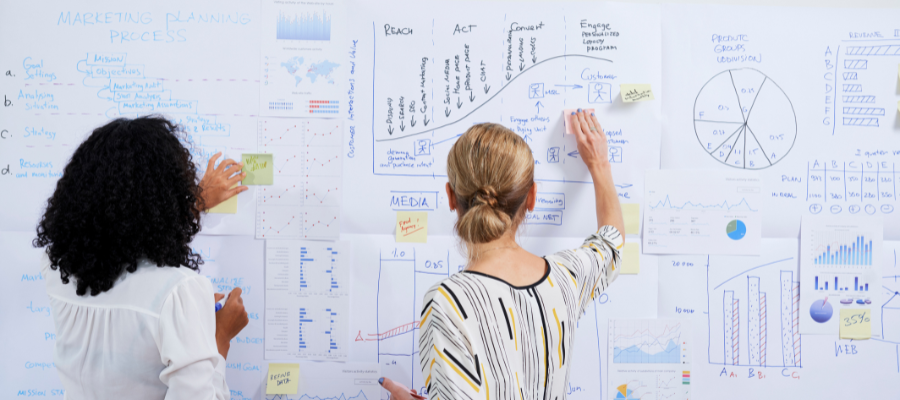 Two women brainstorming in front of a wall filled with charts and diagrams, representing strategic leadership planning.