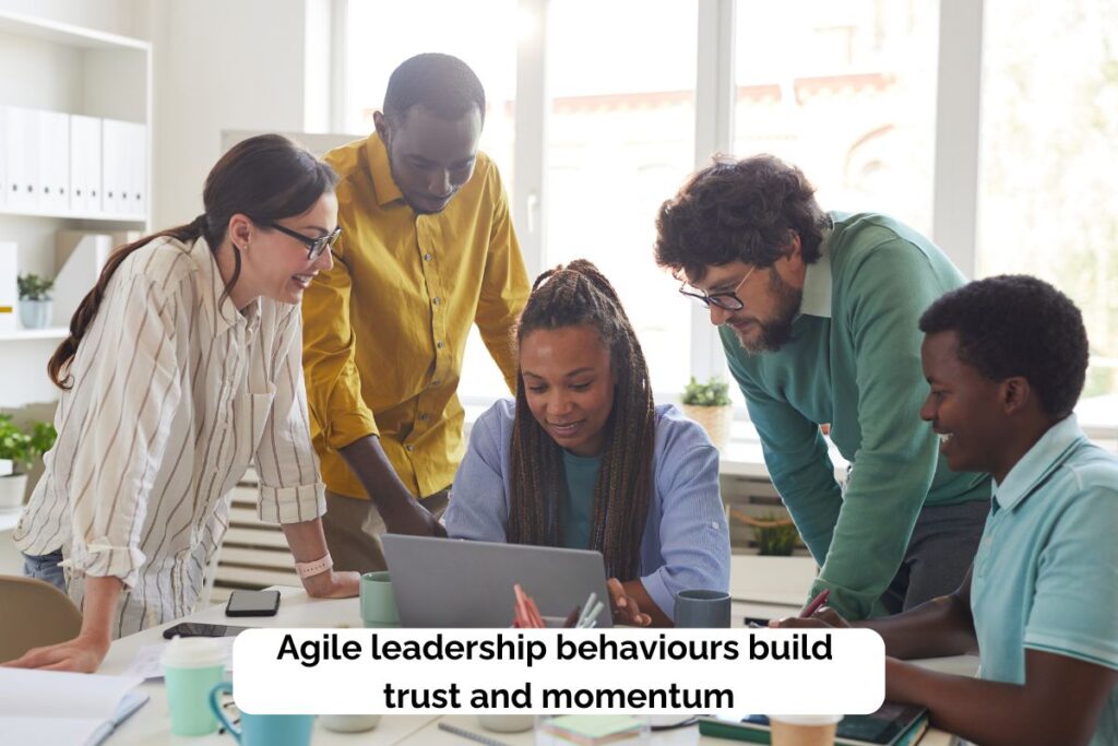 A diverse team gathered around a laptop smiling and collaborating. Text reads “Agile leadership behaviours build trust and momentum.”