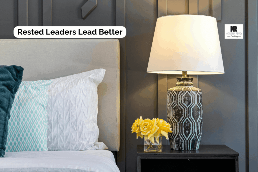 Close-up of a neatly made bed with pillows and a bedside table holding a decorative lamp and yellow flowers. Text overlay reads: ‘Rested Leaders Lead Better.’