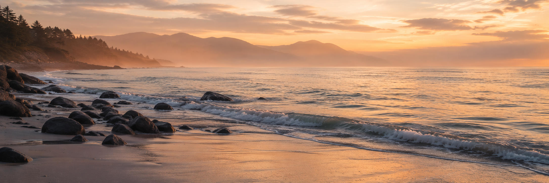 Gentle waves washing onto a rocky beach at sunset with distant mountains fading into warm coastal mist.